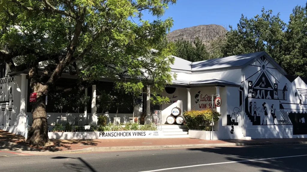 Franschhoek wine shop on the main street, with white Cape Dutch-style buildings, shaded trees, a wall mural, and mountains in the background.