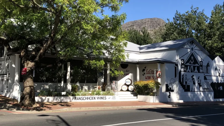 Franschhoek wine shop on the main street, with white Cape Dutch-style buildings, shaded trees, a wall mural, and mountains in the background.