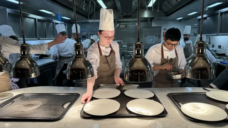 Chefs plating at the pass in a fine dining restaurant kitchen, white plates lined up under heat lamps.