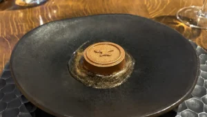 Erst, Manchester, The Flatbread We Did Not Order – The Anvil dessert at L'Enclume, Cartmel: a copper-coloured caramel mousse disc stamped with an anvil, in a gold-flecked pool on a black plate
